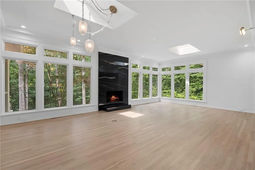Spacious living area featuring extensive window arrays, light wood-finish flooring, a black fireplace with a stone surround, and an overhead skylight