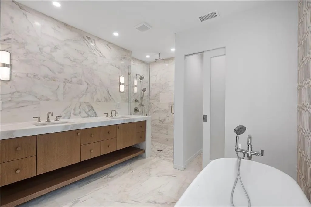 Contemporary bathroom featuring a freestanding soaking tub with a handheld shower, a dual vanity with wood-finish cabinetry and integrated sinks, a glass-enclosed shower, and extensive marble-style wall and floor tiling