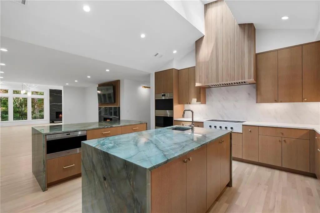 Open-concept kitchen featuring two islands with unique stone countertops, light wood-finish cabinetry, a slatted wood range hood, integrated appliances, and light wood-finish flooring