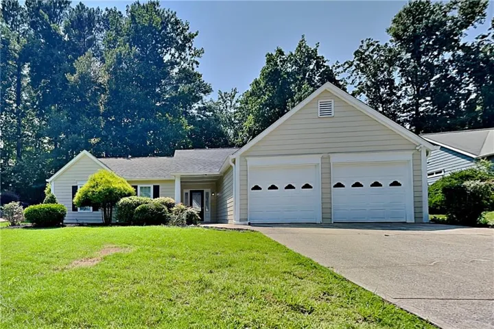 Ranch-style residence featuring light-toned siding, a two-car garage with decorative paneling, a paved driveway, and mature landscaping