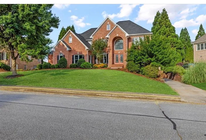 Brick facade residence featuring a prominent arched window, black shuttered windows, a well-maintained lawn, mature trees, and a concrete driveway