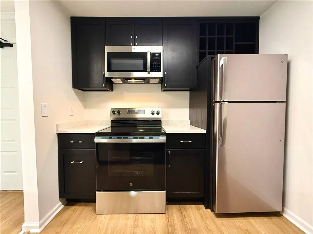 Kitchen with dark cabinetry, appliances with stainless steel finishes, light wood-style flooring, and light stone counters