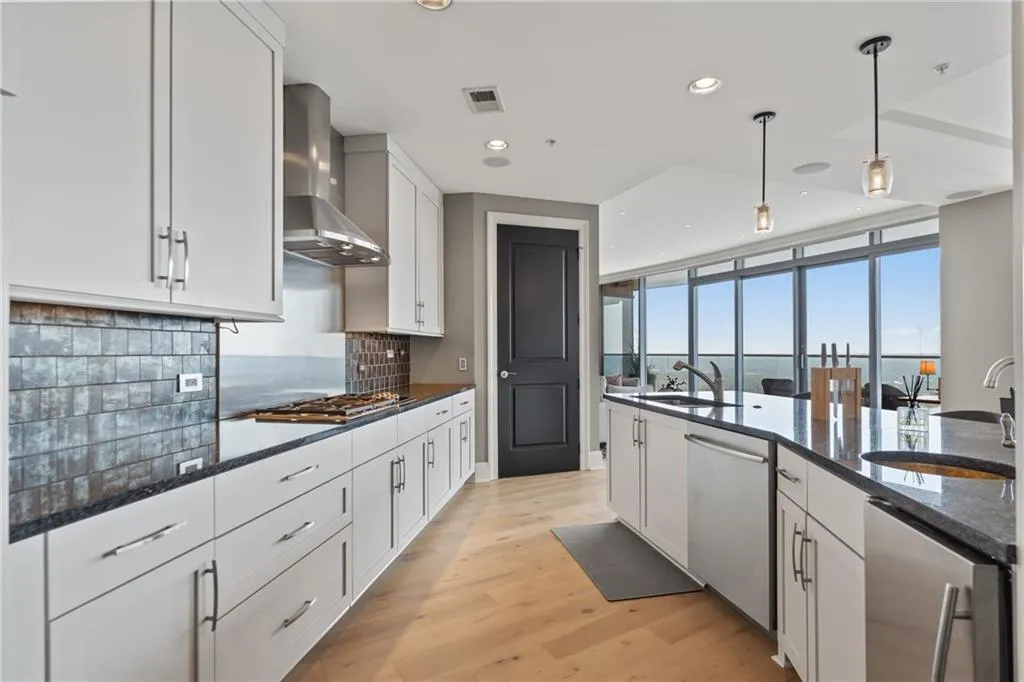 Kitchen with dark stone counters, decorative light fixtures, white cabinetry, wall chimney exhaust hood, and light wood-style flooring