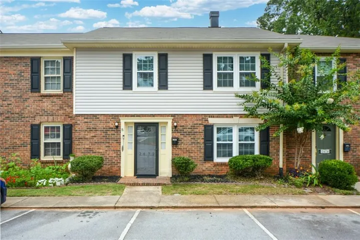 Two-story townhouse with a brick and siding exterior, black window shutters, a light-colored main entry door with a screen door, and foundational shrubs