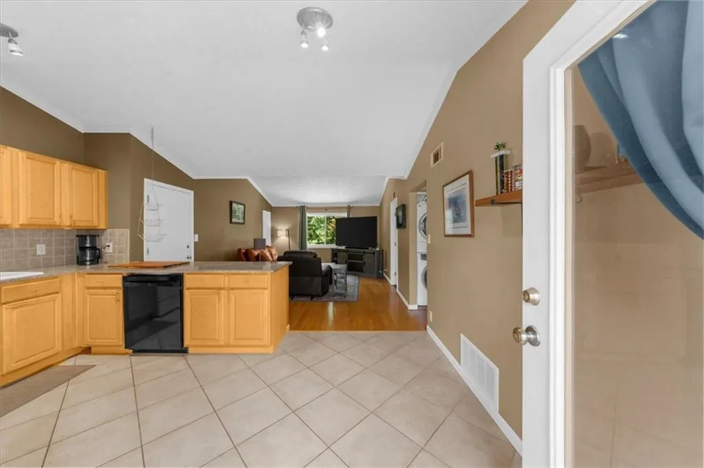 Kitchen featuring dishwasher, stacked washing machine and dryer, light brown cabinetry, vaulted ceiling, and decorative backsplash