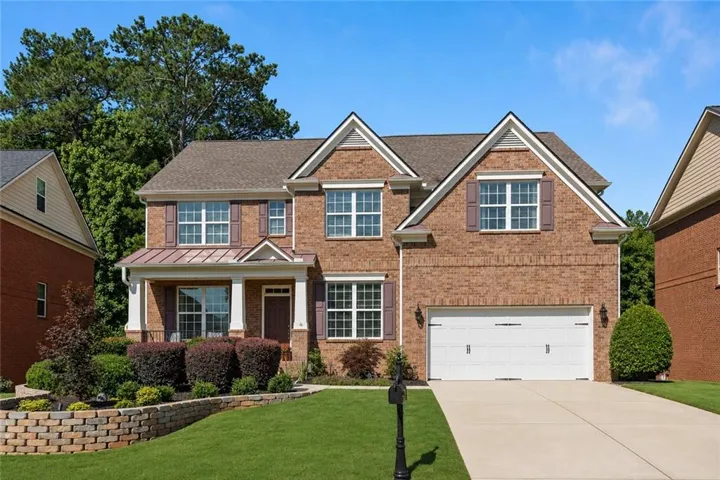 Brick-front residence featuring a covered entry porch with white columns, a two-car garage, mature landscaping, and an elevated front yard with a retaining wall