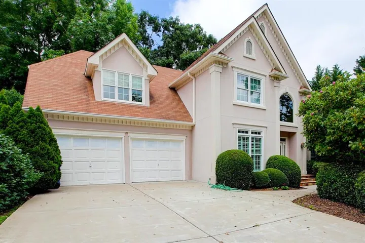 Stucco exterior with a two-car garage, dormer window, arched entry, and mature landscaping