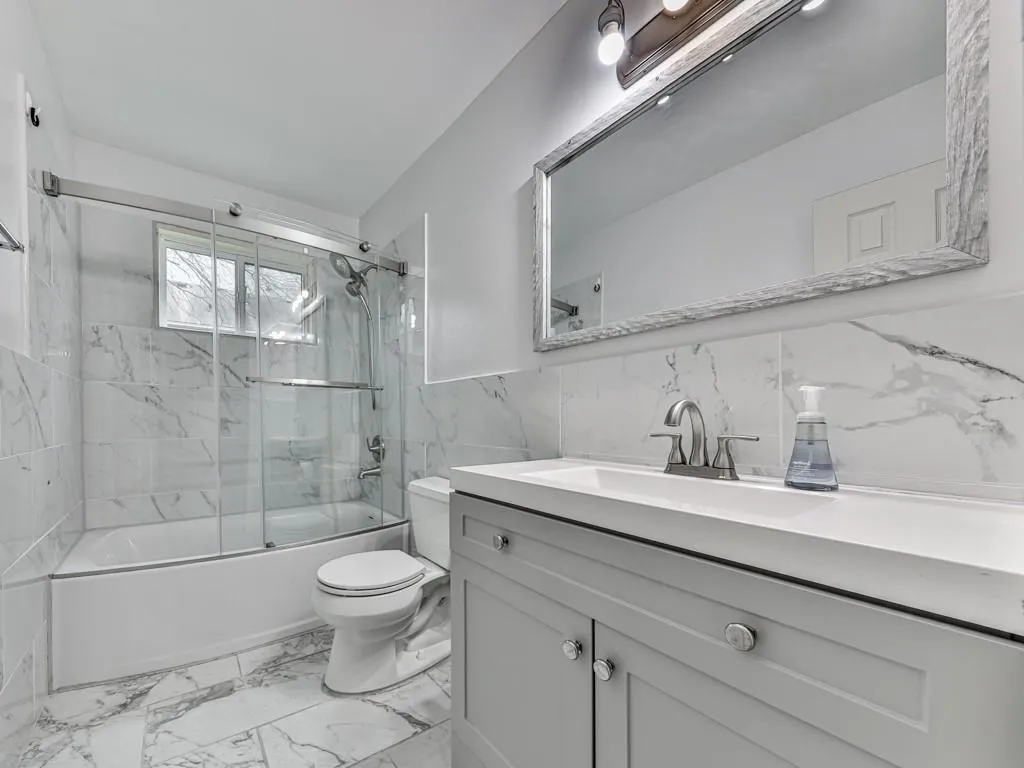 Bathroom featuring enclosed tub / shower combo, light marble finish floors, vanity, and tile walls