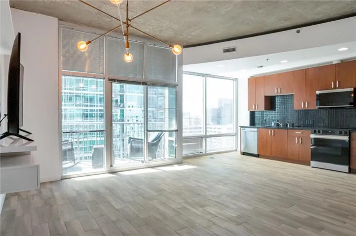 Expansive windows with balcony access, wood-finish flooring, exposed concrete ceiling, contemporary chandelier, and a kitchen featuring wood cabinetry, stainless steel appliances, and a tiled backsplash