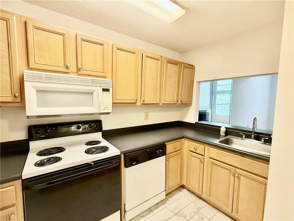 Kitchen with light brown cabinets, white appliances, light marble finish flooring, dark countertops, and a textured ceiling