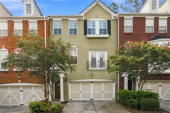 Charming townhome featuring a cream-colored facade with black and white garage doors, flanked by mature landscaping and shrubs
