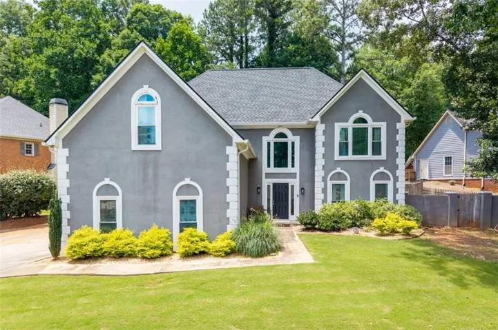 Gray stucco exterior featuring white trim accents, arched windows, a dark paneled front door, a gabled roofline, and established front landscaping