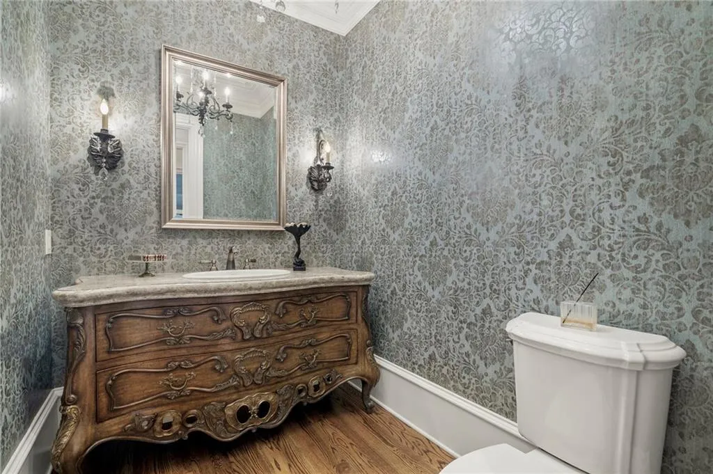 Decorative damask pattern wallpaper, a carved wood-finish vanity with a stone countertop, an undermount sink, and wall-mounted sconces