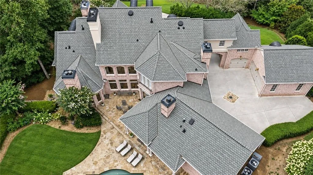 Expansive brick exterior with a multi-level shingle roof, a large stone patio, an inground pool, and mature landscaping
