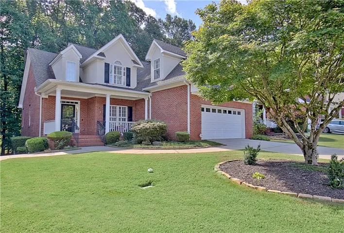 Brick exterior with dormer windows, covered front porch, attached garage, and mature landscaping