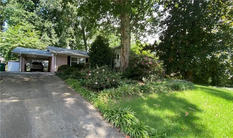 Single-story residence featuring a carport, mature trees, established landscaping, and a sloped front lawn