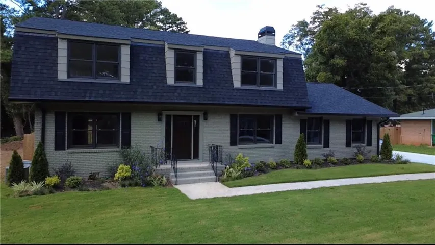 Mansard-style roofline with dormer windows, painted brick exterior, dark window shutters, concrete walkway, and a manicured lawn