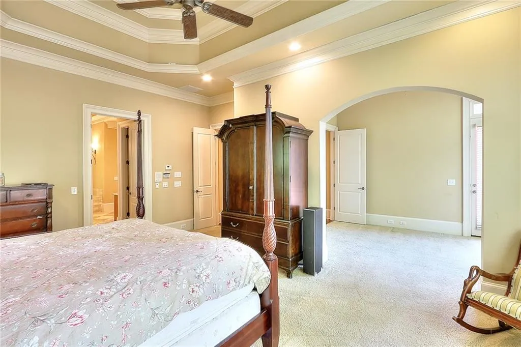 Carpeted bedroom with a raised ceiling, ornamental molding, ensuite bath, and ceiling fan