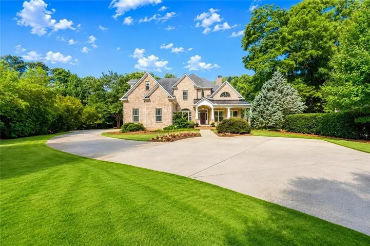 Brick-clad residence featuring a covered front porch with columns, a curved driveway, mature trees, and manicured landscaping