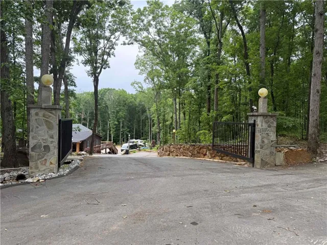 Gated asphalt driveway with stone pillars, featuring a wooded backdrop and visible building structures