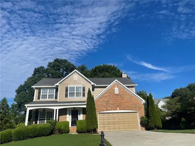 Two-story residence featuring a combination of brick and siding, an integrated two-car garage, a covered front porch with white columns, a paved driveway, and mature landscaping