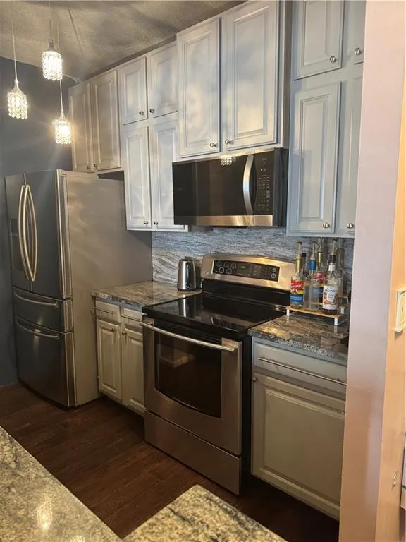 Kitchen featuring appliances with stainless steel finishes, backsplash, dark stone countertops, hanging light fixtures, and dark wood-style floors