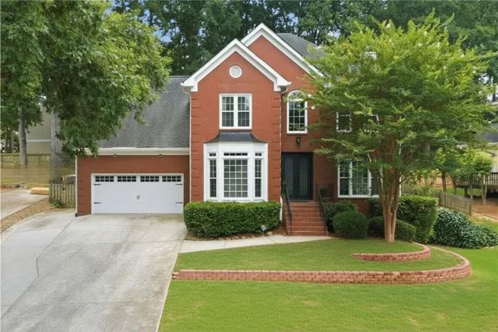 Traditional brick exterior featuring white trim accents, a prominent bay window, a two-car garage, manicured lawn with brick retaining walls, and mature landscaping