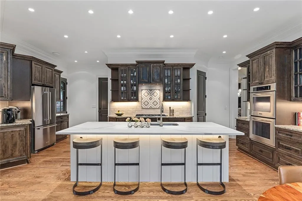 Kitchen featuring glass insert cabinets, dark brown cabinets, recessed lighting, a center island with sink, and light stone counters