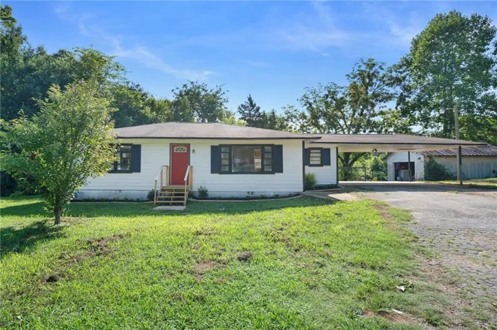 White siding residence featuring a red entry door, dark shutters, a front porch with natural wood steps, a gravel driveway, and an attached carport