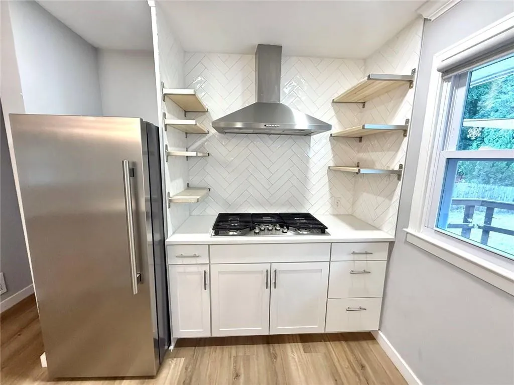 Kitchen featuring stainless steel appliances, open shelves, wall chimney exhaust hood, tasteful backsplash, and light countertops