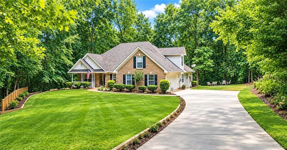Brick exterior with white trim, covered front porch, attached garage, curved driveway, and lush landscaping