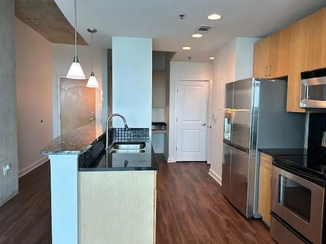 Kitchen with appliances with stainless steel finishes, dark wood-style flooring, dark stone counters, and decorative light fixtures