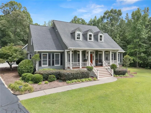 Traditional-style home featuring a covered front porch with white railings, a prominent red front door, multiple dormer windows, a dark shingle roof, and a mature tree-lined background