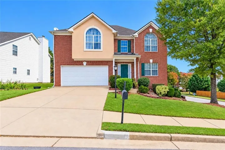 Brick exterior with stucco accents, arched windows, attached garage, well-maintained landscaping, and mature tree