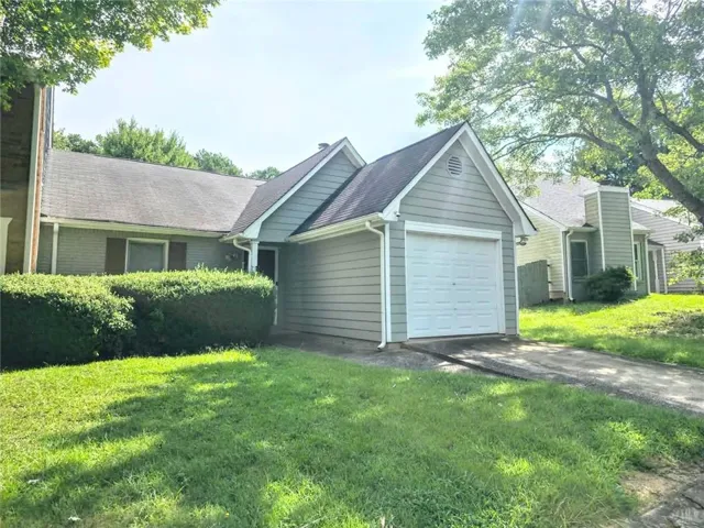 Exterior featuring light green siding, an attached garage, a paved driveway, and mature landscaping