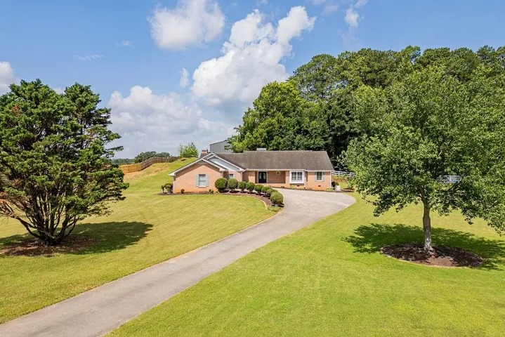 Brick exterior residence with a paved driveway, expansive green lawn, mature trees, and natural landscaping