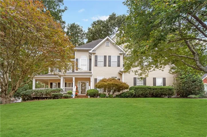 Traditional two-story residence featuring a covered front porch, light-toned siding, dark window shutters, established trees, and a spacious front lawn