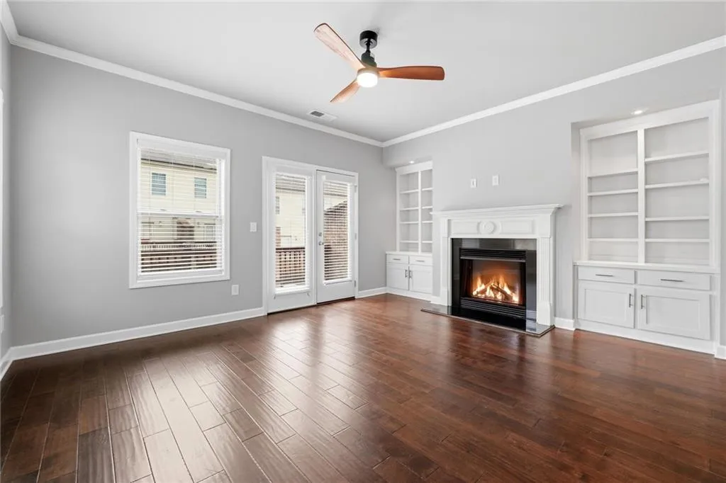 Unfurnished living room with a ceiling fan, dark wood-type flooring, crown molding, a glass covered fireplace, and built in features