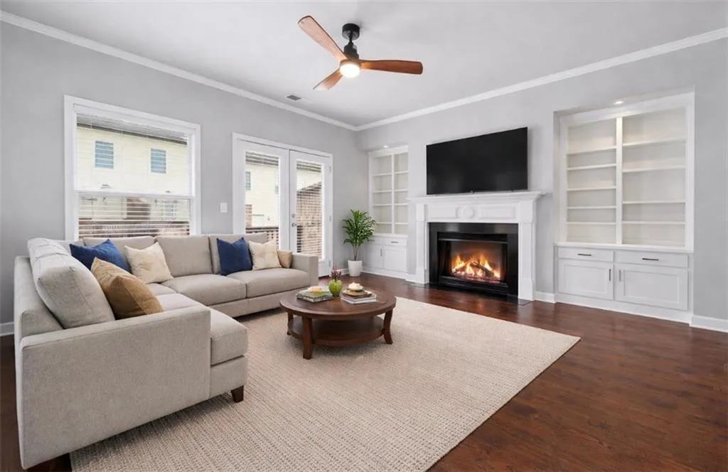 Living area with ornamental molding, dark wood-type flooring, built in shelves, a fireplace with flush hearth, and ceiling fan