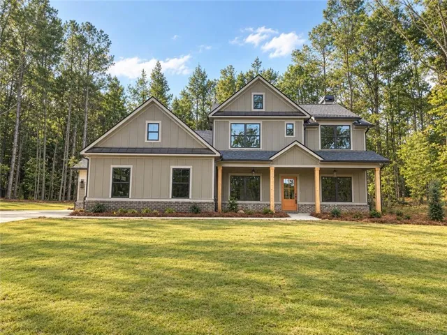 Two-story residence featuring a covered front porch with natural wood columns, board and batten siding, dark framed windows, and a mature treeline backdrop