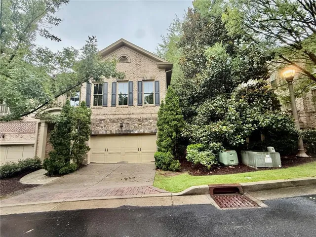 Brick façade home featuring a two-car garage, shuttered windows, a curved driveway, and mature landscaping