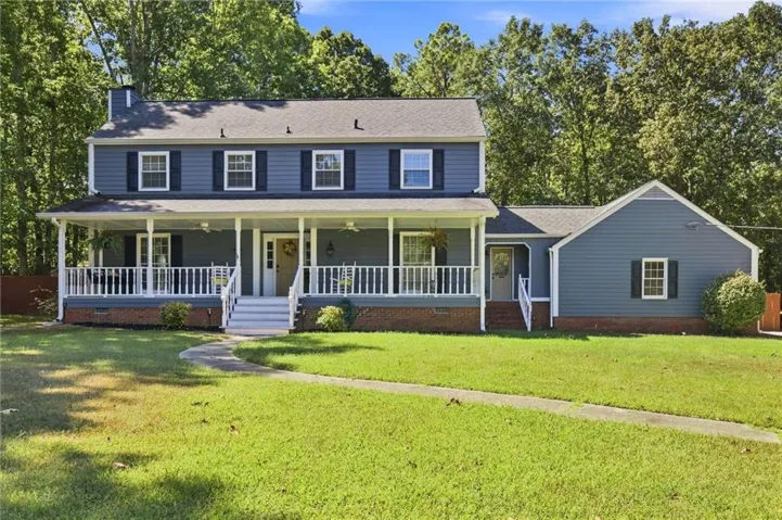 Two-story residence featuring blue siding, white trim, a covered front porch, an expansive lawn, and mature trees as a backdrop