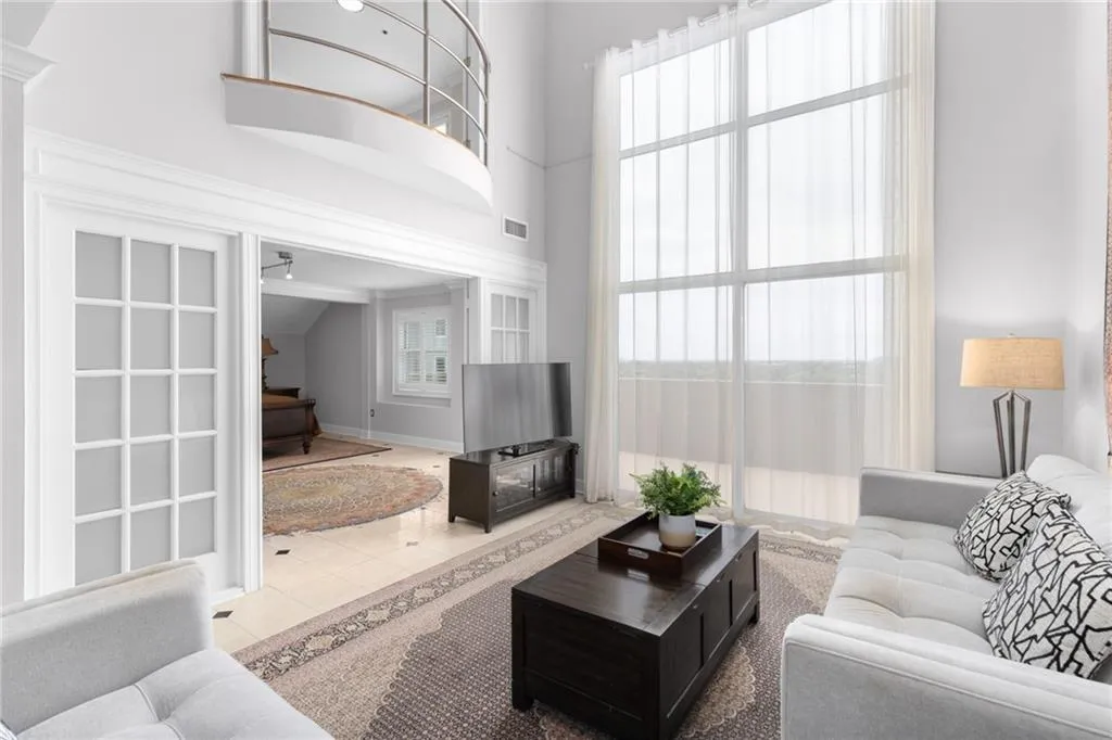 Two-story living area featuring floor-to-ceiling windows with sheer drapery, a second-story balcony with a curved railing, and French doors opening to an additional room