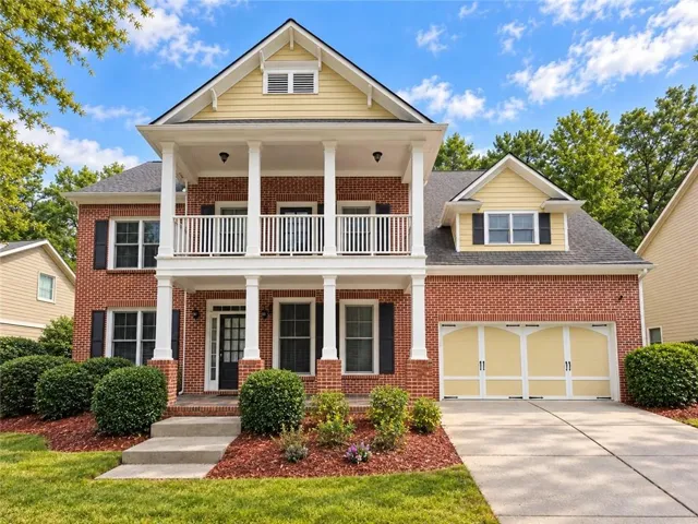 Brick exterior with yellow siding accents, a two-car garage, a covered porch with white columns, a second-story balcony, and mature landscaping