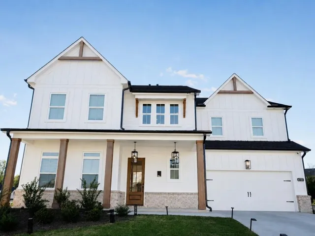 White board and batten exterior with wood-tone accents, covered porch with substantial columns, dark wood-finish front door with glass panels, integrated two-car garage, and exterior lighting fixtures