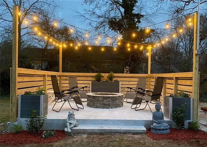 Expansive patio featuring a stone-clad fire pit, slatted wood privacy fencing, string lighting, and raised planter boxes
