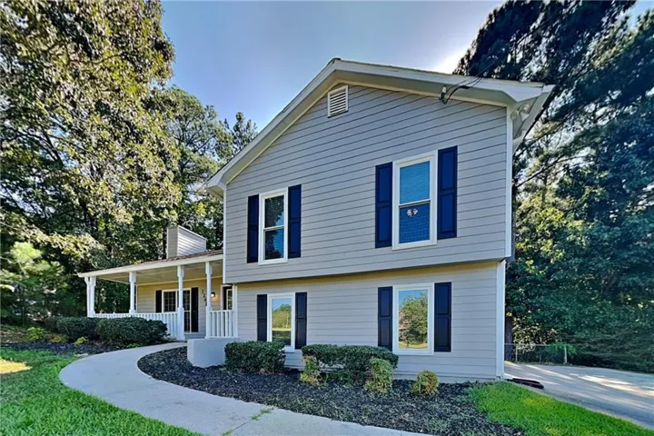 Two-story residence featuring light gray siding with dark blue shutters, a covered porch with white columns, a curving concrete walkway, and established landscaping