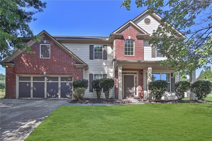 Welcoming two-story brick Colonial featuring a four-car garage with paneled doors, manicured lawn with mature landscaping, and white trim accents