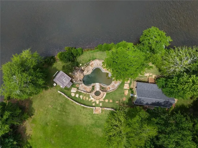 Waterfront property featuring a freeform pool with integrated spa, stone hardscaping, natural rock retaining walls, and a separate structure with a dark shingle roof