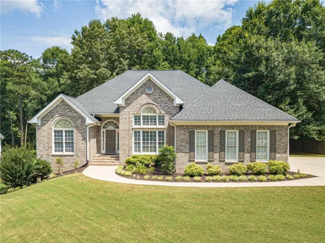 Traditional brick residence featuring a dark gray shingle roof, arched transom windows, dark shutters, a concrete walkway, and a manicured lawn with established landscaping